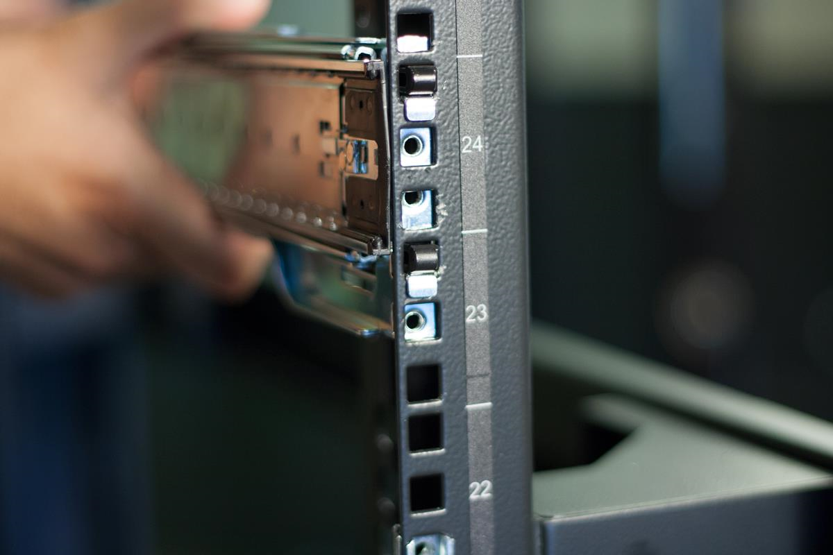 Close-up photo of a technician pressing a server rail into a rack column; shows a hand holding the rail and the numbered mounting holes on the rack